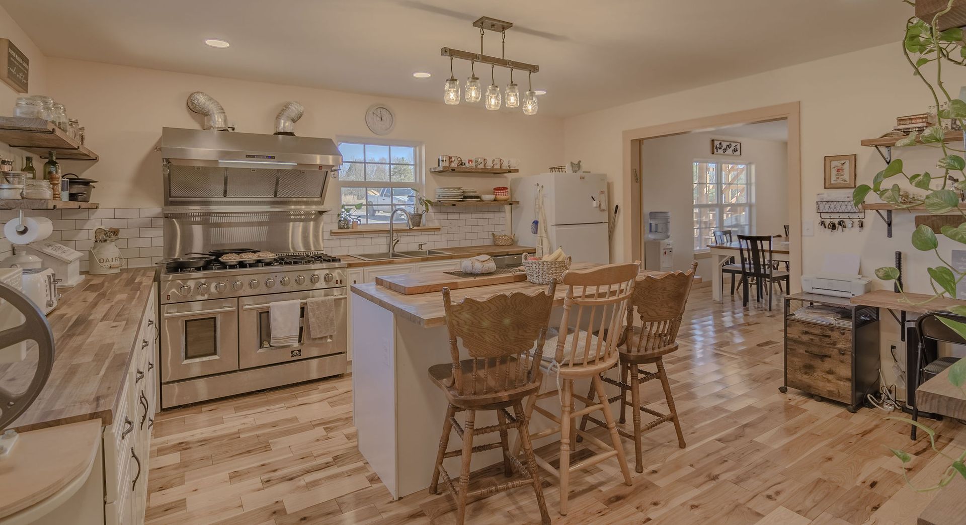A rustic kitchen with a large stainless steel range, a central island with three wooden chairs, and light wood flooring.