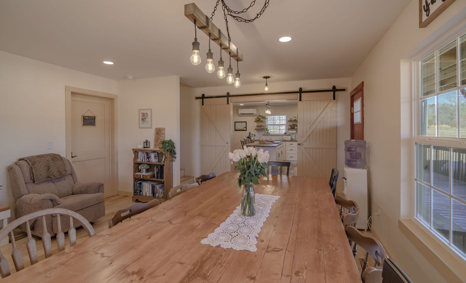 Dining room featuring a long wooden table, farmhouse chandelier, armchair, and barn doors opening to a kitchen.