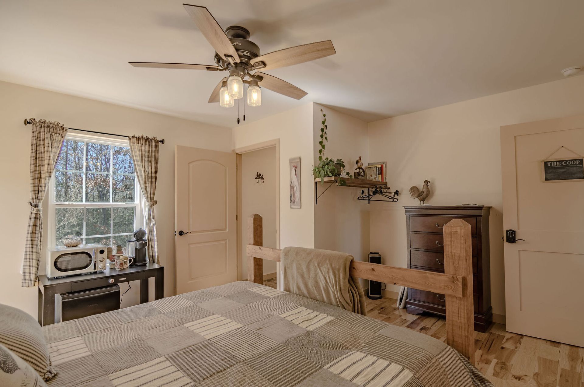 A cozy bedroom with a quilted bed, a ceiling fan, a window with plaid curtains, and a wooden dresser in a neutral room.