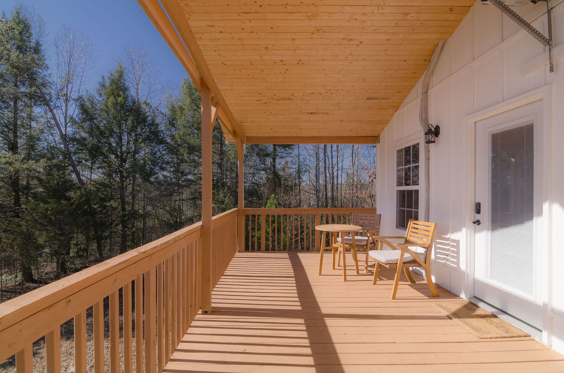 A wooden porch with chairs and a small table overlooking a forest, attached to a white building with a glass door.