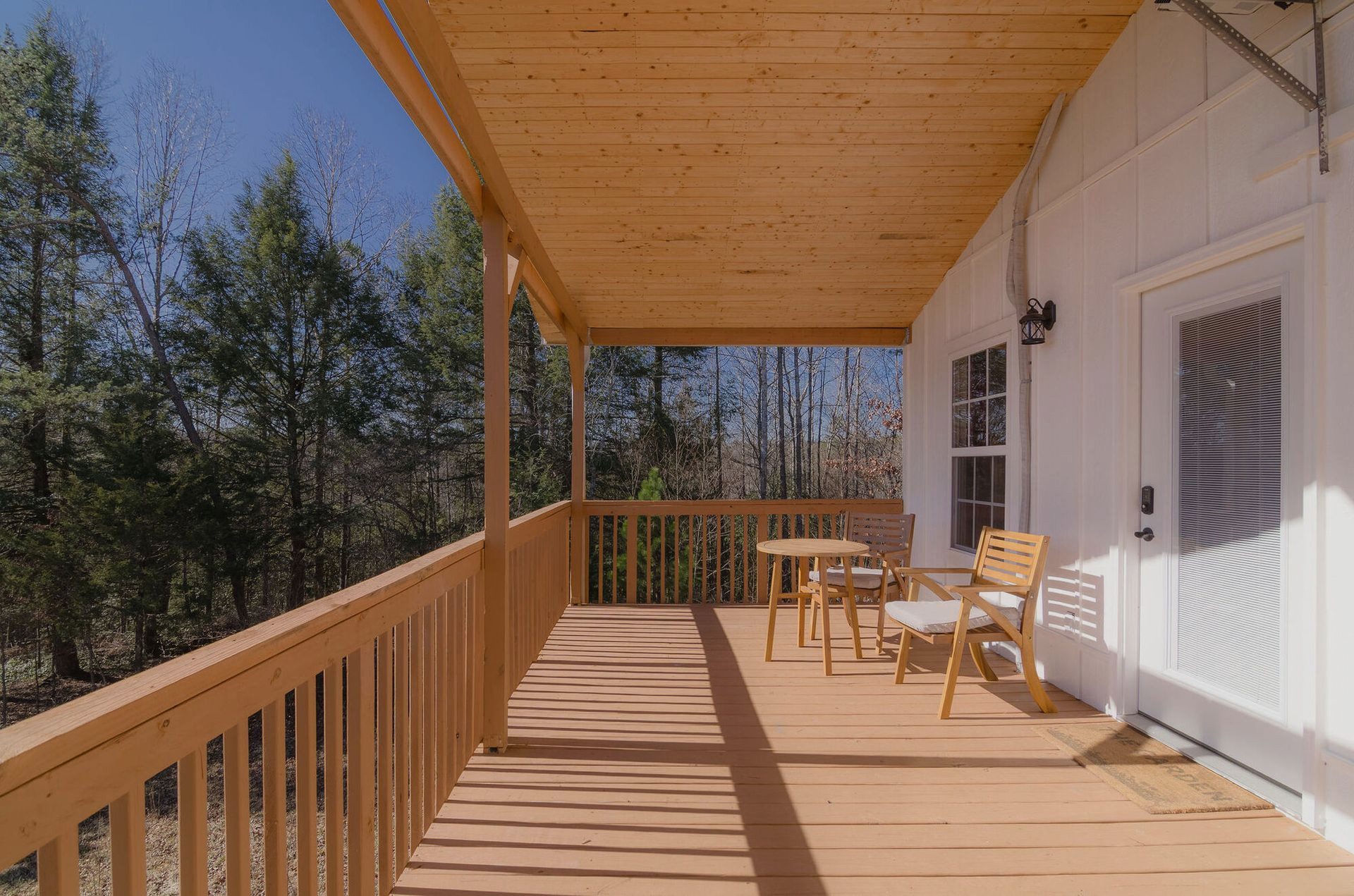 A sunlit wooden porch featuring a small round table and two chairs, overlooking a wooded forest area.
