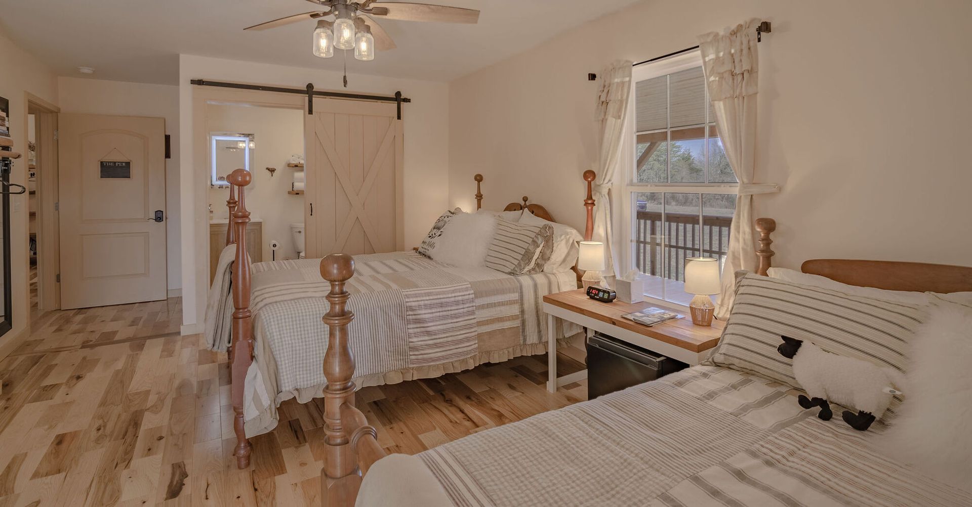 A cozy bedroom with two wooden beds, white bedding, a sliding barn door, and a window with lace curtains.