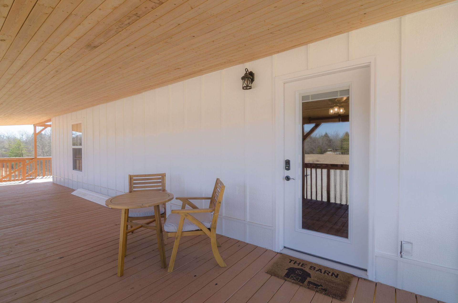 A covered wooden deck features a small round table with two chairs, white walls, a glass-paneled door, and a door mat.