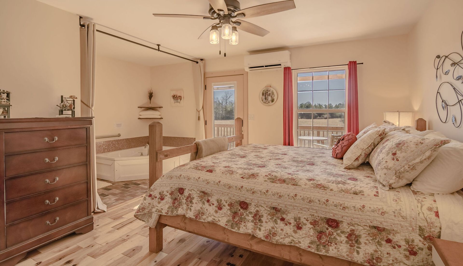 A cozy bedroom featuring a floral quilt bed, wooden dresser, ceiling fan, and an open bathroom with a jetted tub.