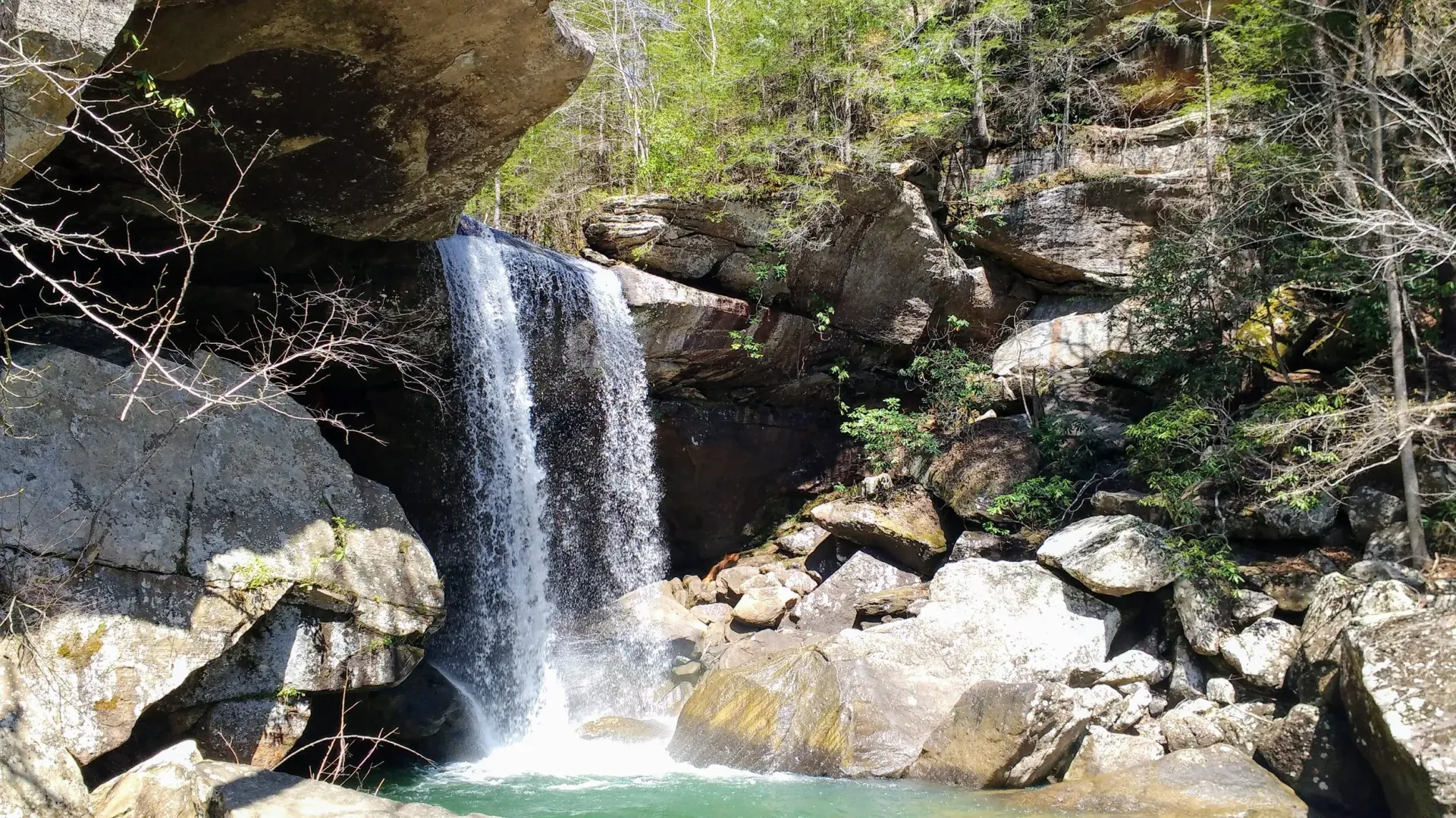 A waterfall cascades over a rocky ledge into a clear turquoise pool, surrounded by rugged boulders and forest foliage.