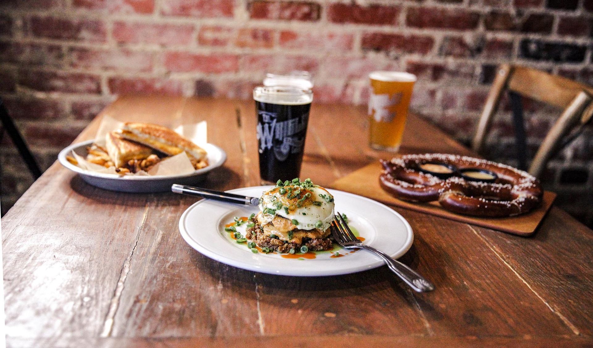 A wooden table with a plate of eggs, a soft pretzel, fries, and two glasses of beer against a brick wall background.