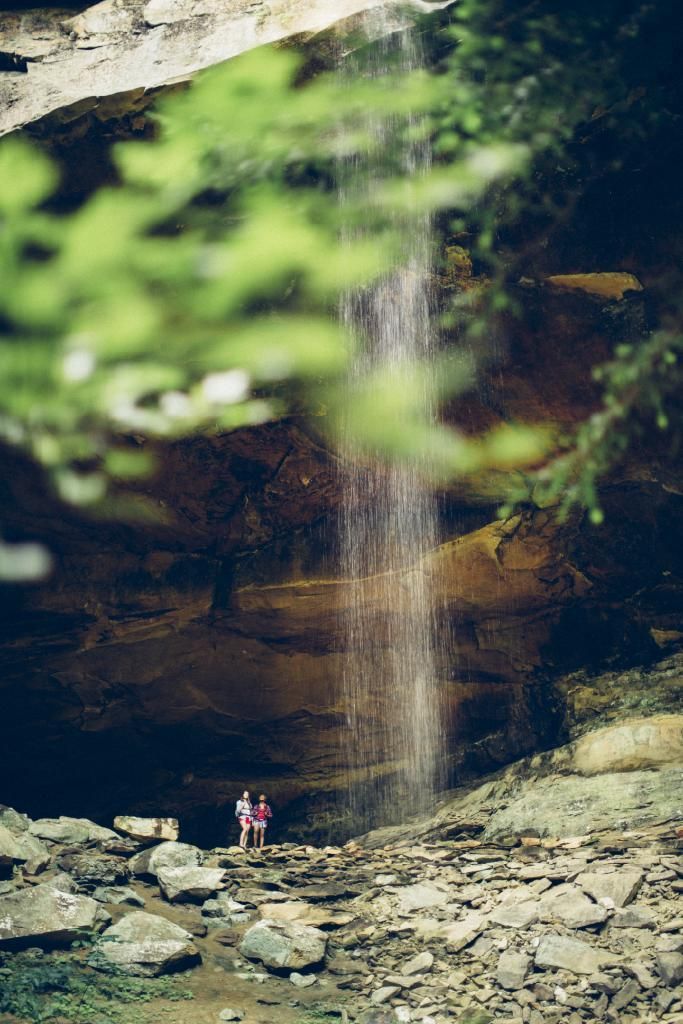 A thin waterfall cascades down a large, layered rock cliff, with two small figures standing near the base.