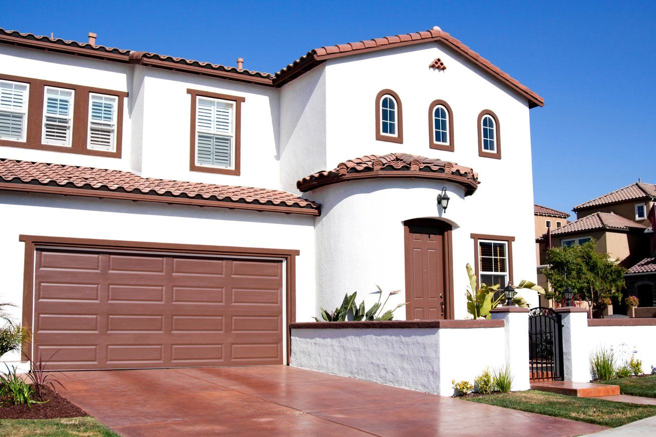 Two-story stucco house with brown garage door, windows, and roof tiles; blue sky.