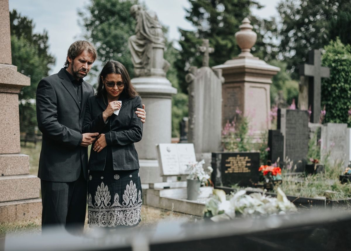 Two mourners stand together at a cemetery, grieving beside a gravestone and flowers.