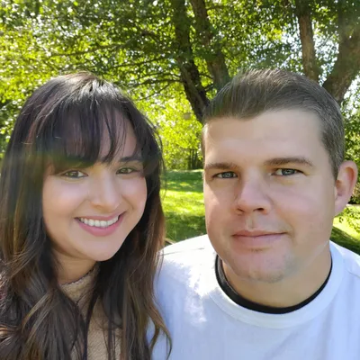 Two people smiling outdoors with green trees in the background