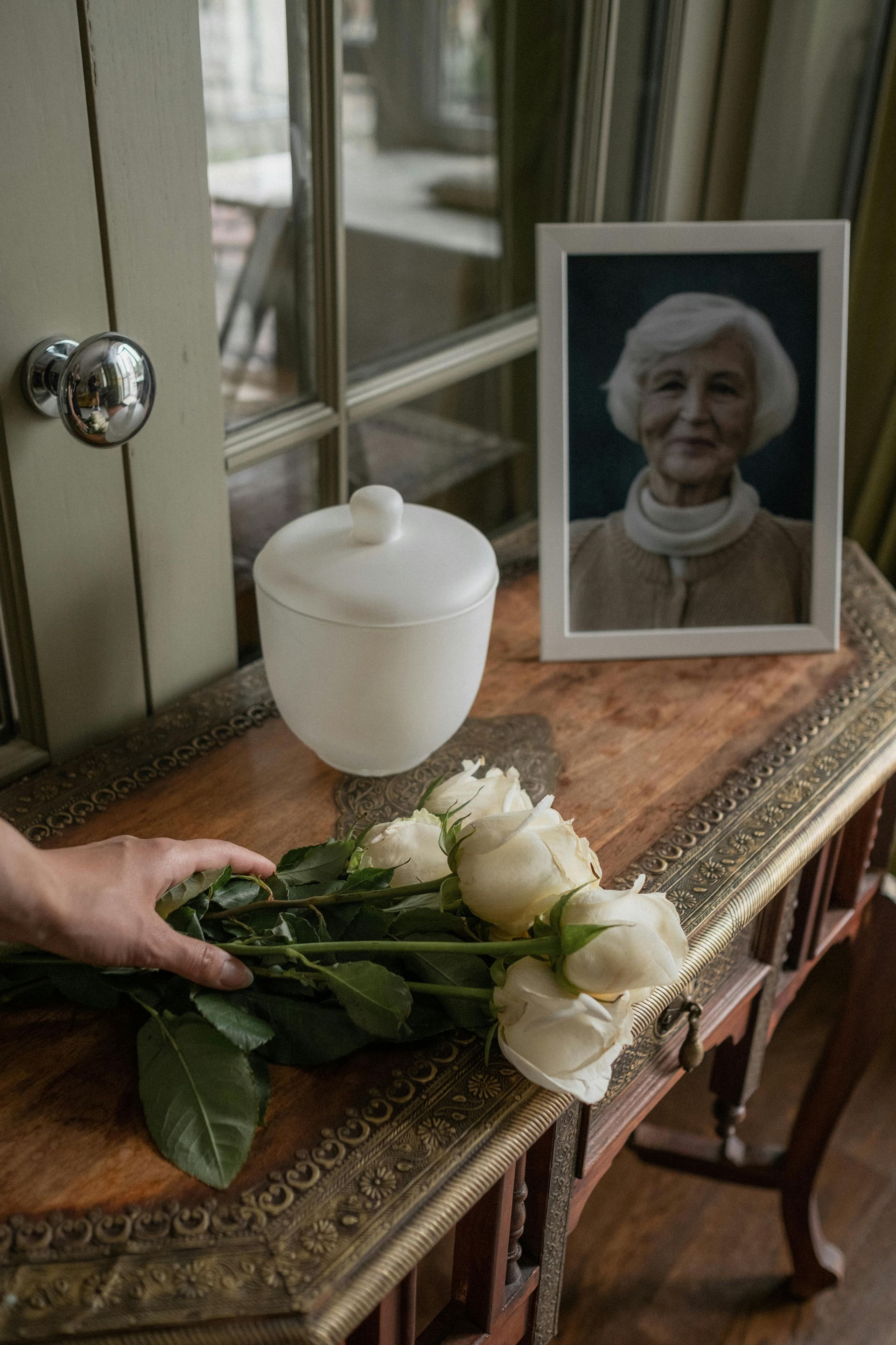 White flowers and a framed portrait on a wooden table, with a candle jar and a hand placing flowers.