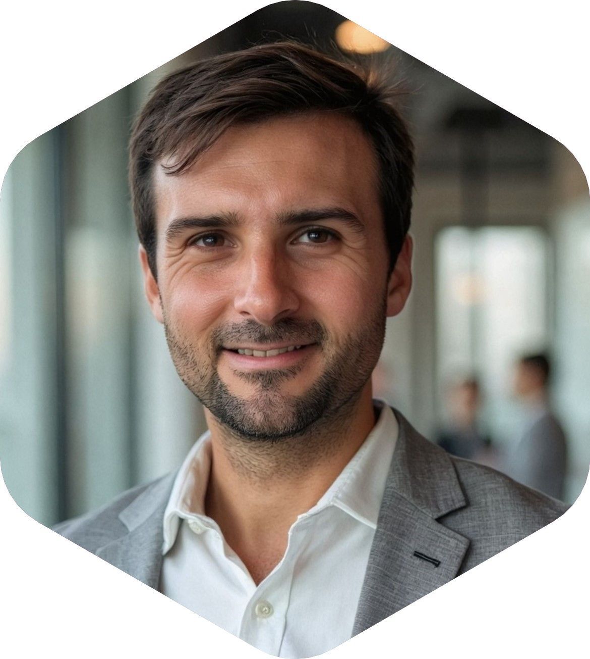 Man in a blazer and open white shirt smiles toward the camera. Blurred office background.