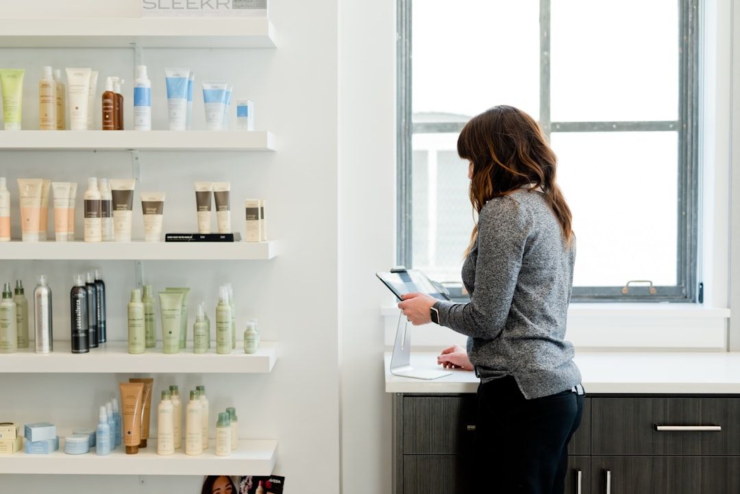 Woman in a salon looking at a tablet next to a shelf filled with skincare products. Sunlight streams in through a window.
