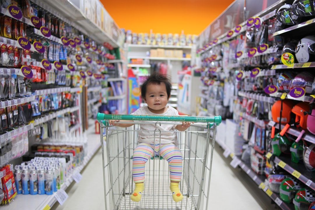 A baby sits in a shopping cart in a store aisle, looking forward with a neutral expression. Shelves with products line both sides.