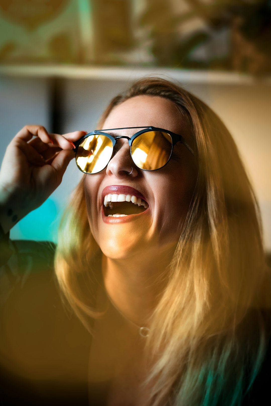 Woman with blonde hair laughing, holding sunglasses up, gold lens reflecting light.