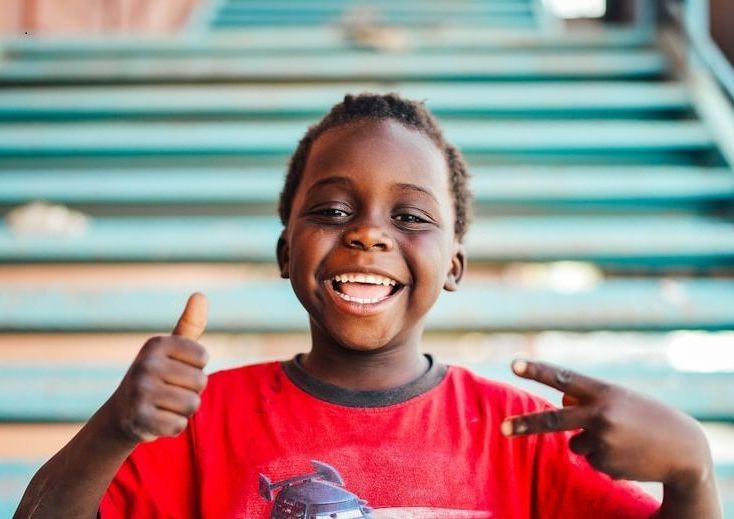 Smiling Black child with thumbs up and peace sign, in front of teal stairs, wearing a red shirt.