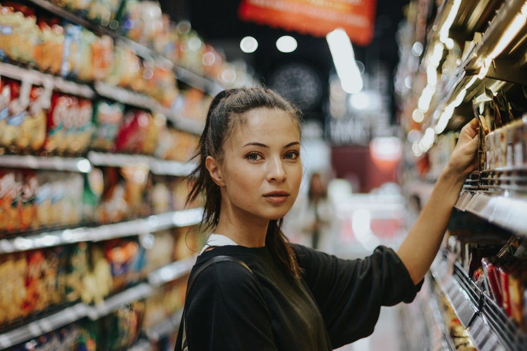 Woman shopping in a grocery store, reaching for items on a shelf. She has dark hair, a neutral expression, and wears a black shirt.