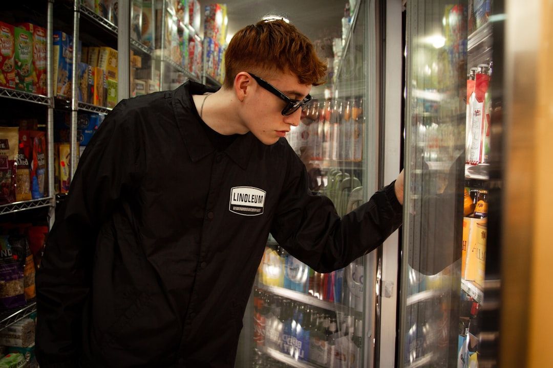 A young man with red hair and sunglasses peers into a refrigerated display case at a convenience store, searching for a product.