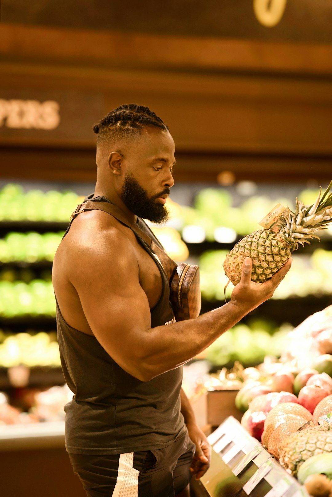 Muscular man in a tank top examines a pineapple in a grocery store produce section. He has a beard and short braids.