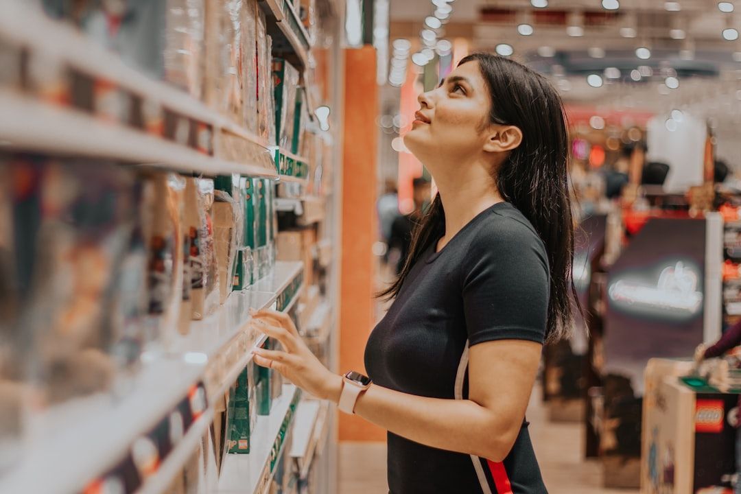 Woman browsing a store shelf, looking upward with a thoughtful expression, in a brightly lit retail environment.