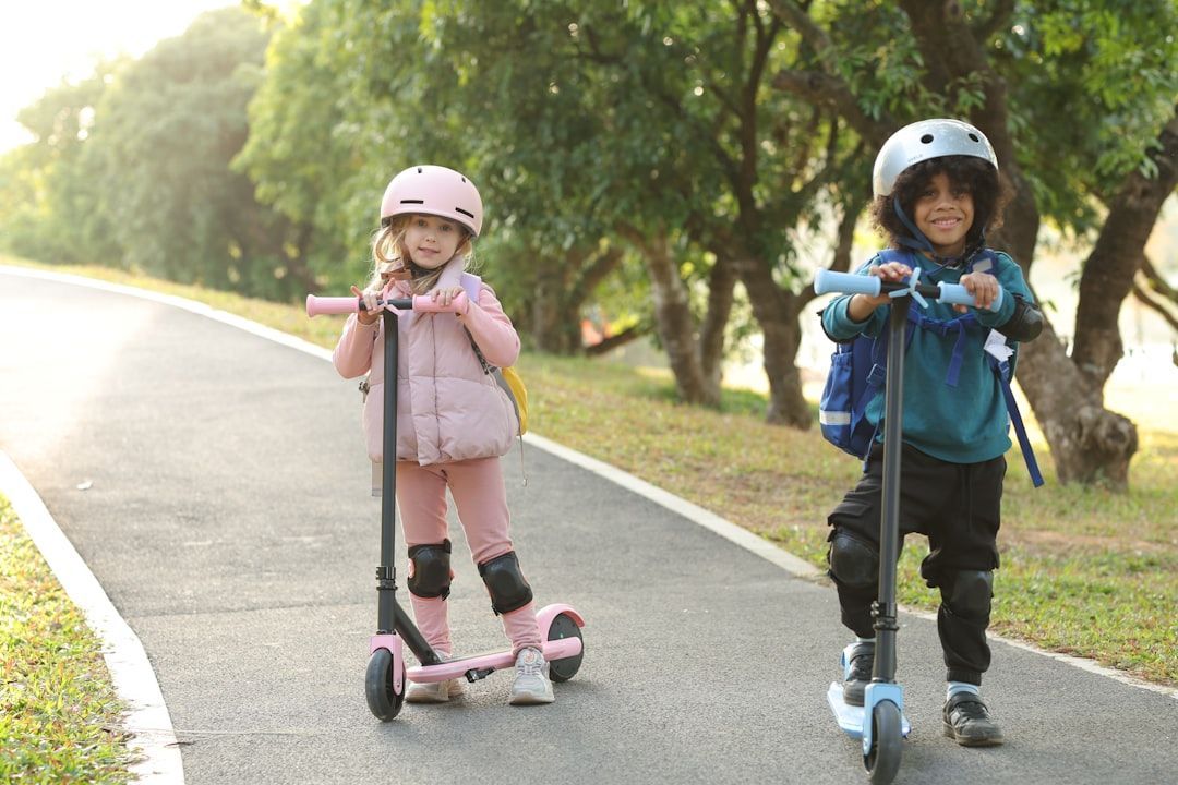 Two children on scooters on a paved path, wearing helmets and pads. One is pink, the other is blue.