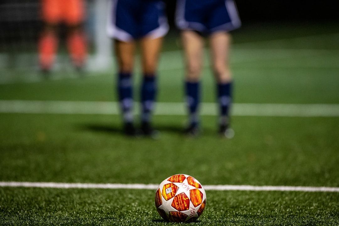 Soccer ball on the field's white line, with two players in blue shorts and a distant player in orange.