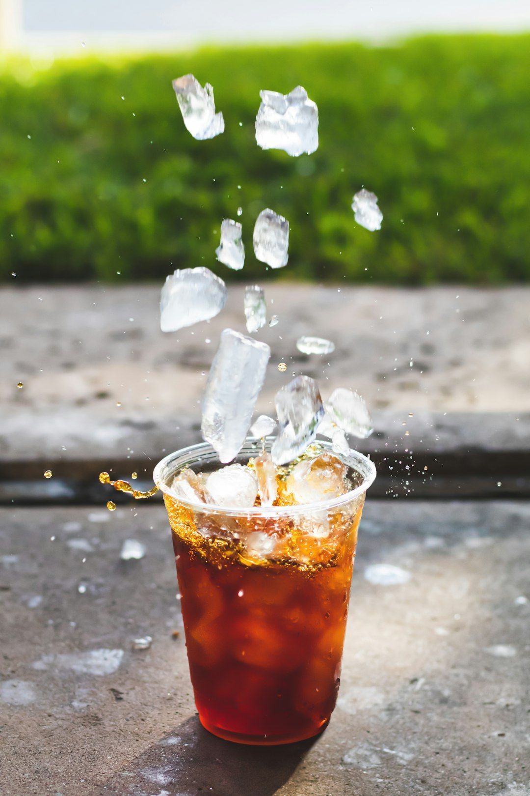 Ice cubes exploding from a clear plastic cup of dark liquid, possibly coffee or cola, on a concrete surface.