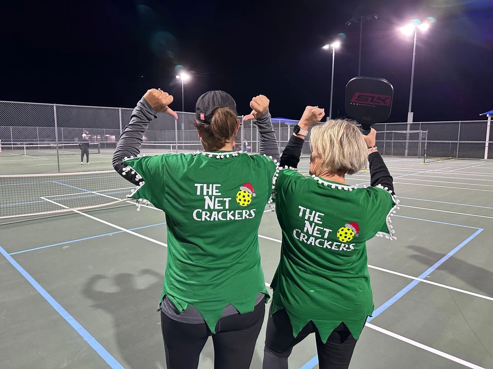 Two women wearing green shirts that say the net crackers are standing on a tennis court.