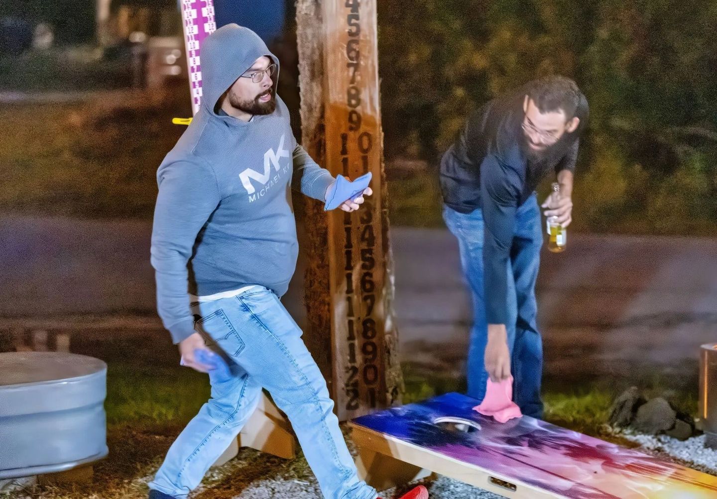 Two men are playing a game of cornhole outside at night.