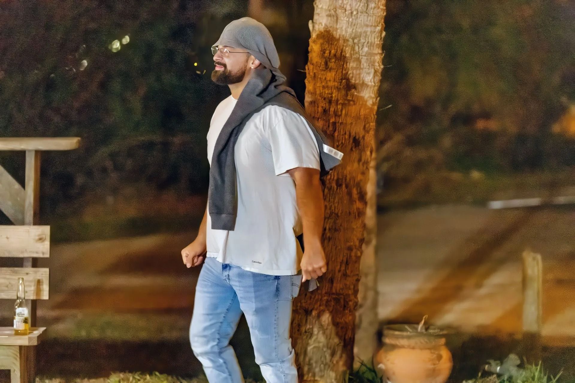 A man in a white shirt and blue jeans is standing next to a tree.