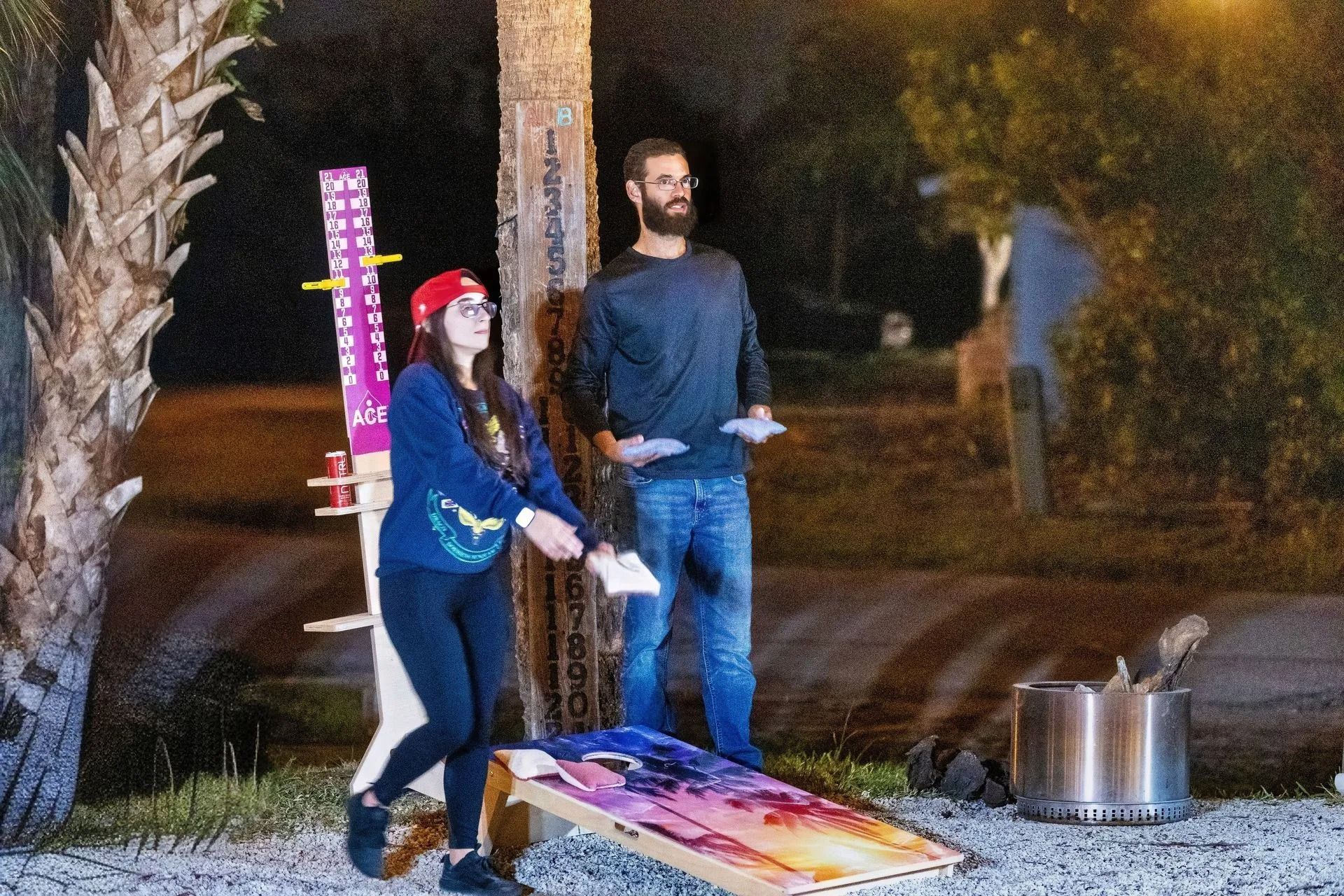 A man and a woman are playing cornhole outside at night.