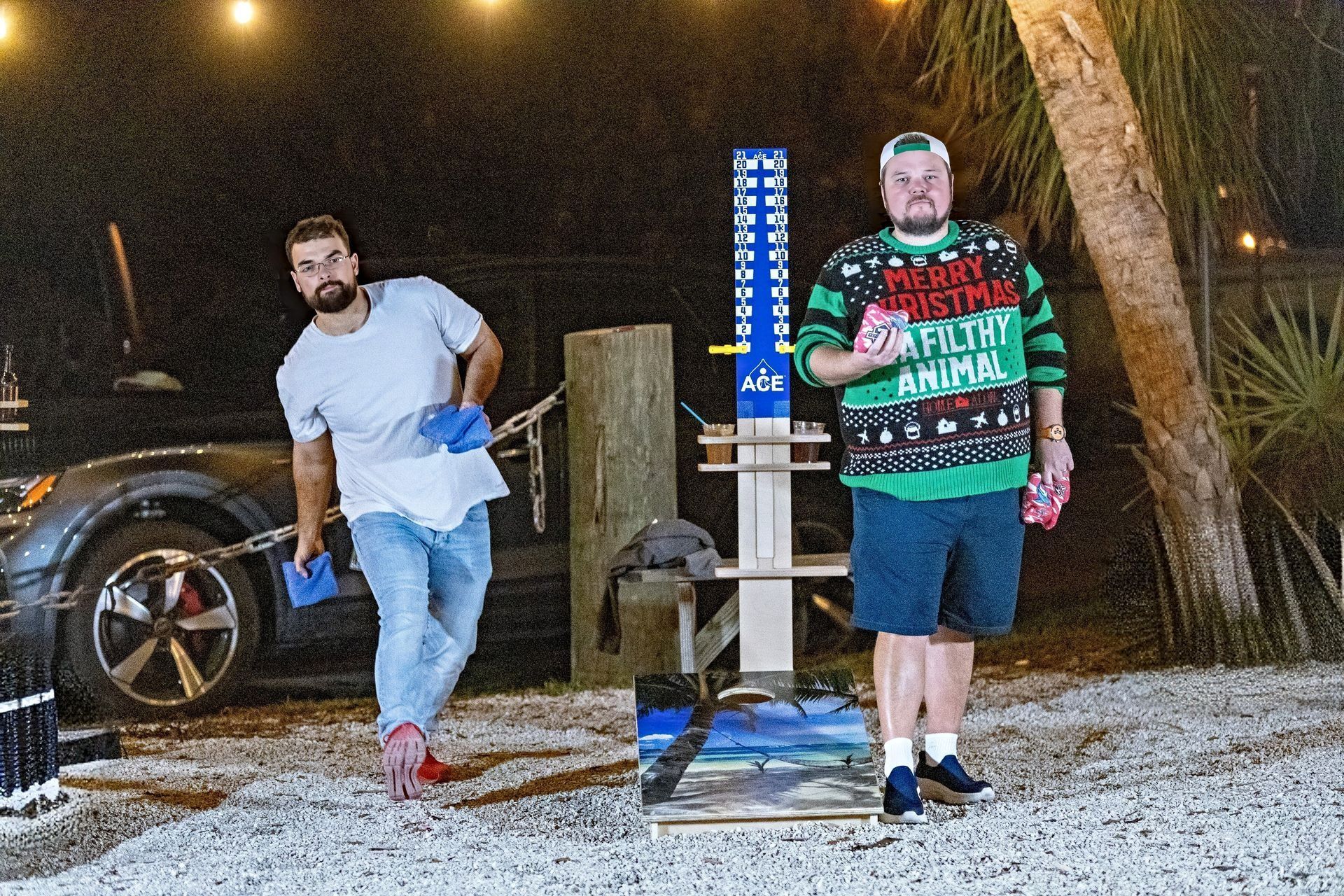 Two men are playing a game of cornhole outside at night . one of the men is wearing a christmas sweater.
