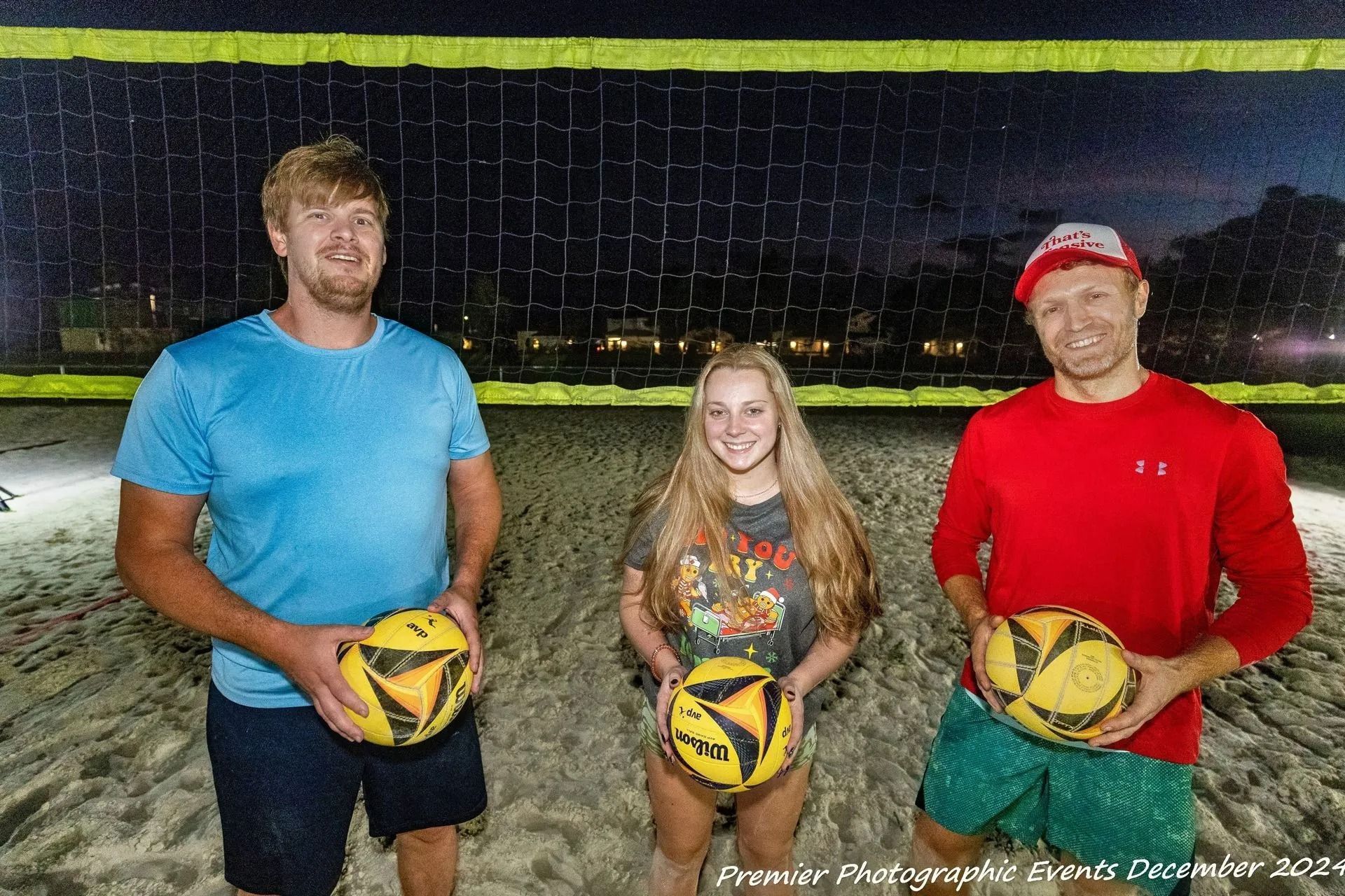 Three people are standing on a beach holding volleyballs.