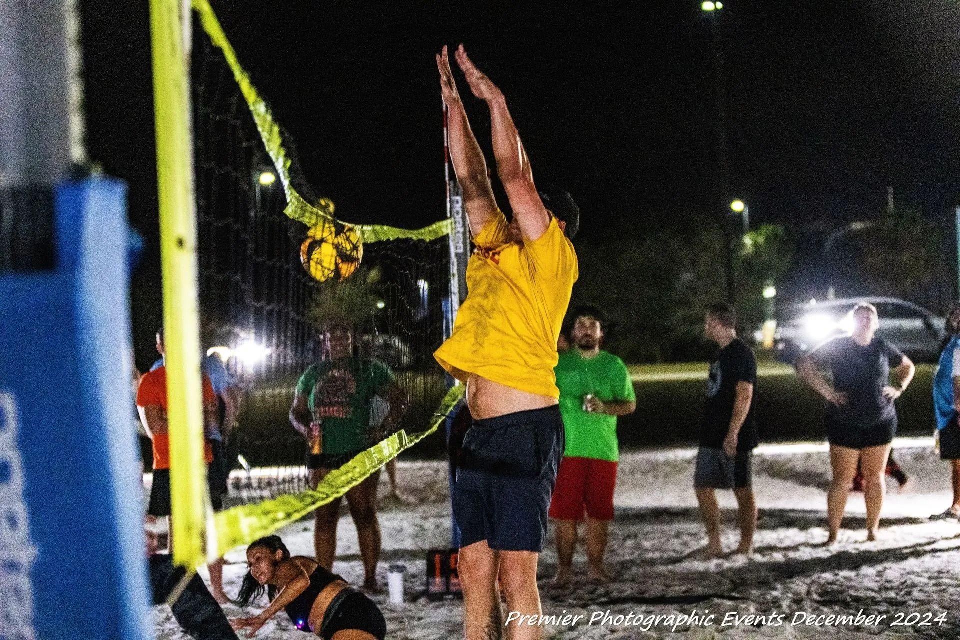 Volleyball player jumps to spike at night; others watch. Yellow net, court, beach setting.