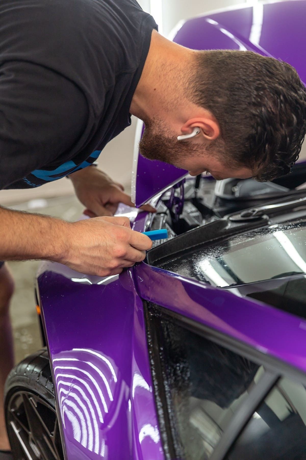 Installer applying a glossy purple vinyl wrap to the fender of a sports car using a squeegee tool, with close attention to detail near the hood and windshield area.