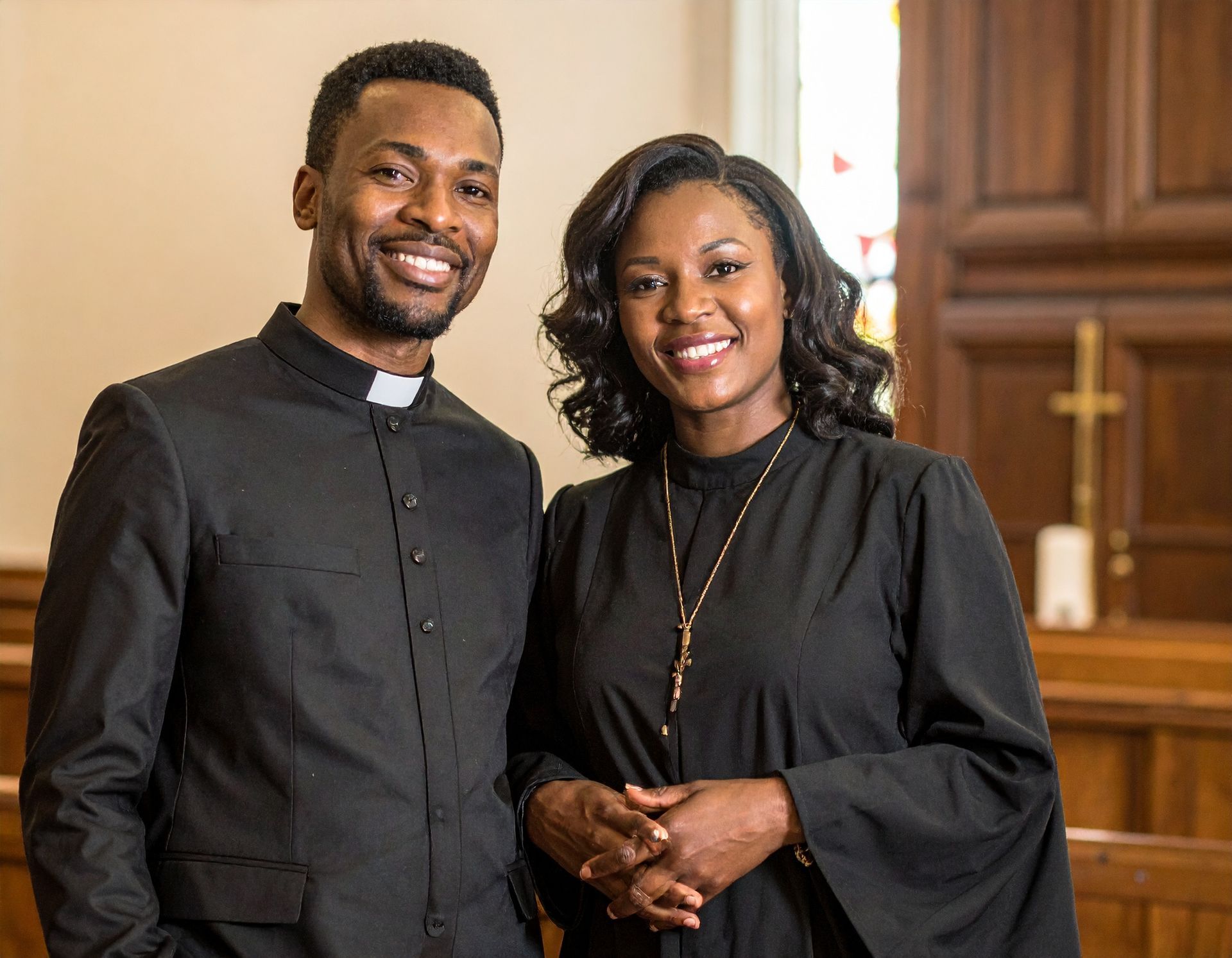 Two people, dressed in clergy attire, smile in a church. Man in black suit; woman in black robe, gold cross necklace.