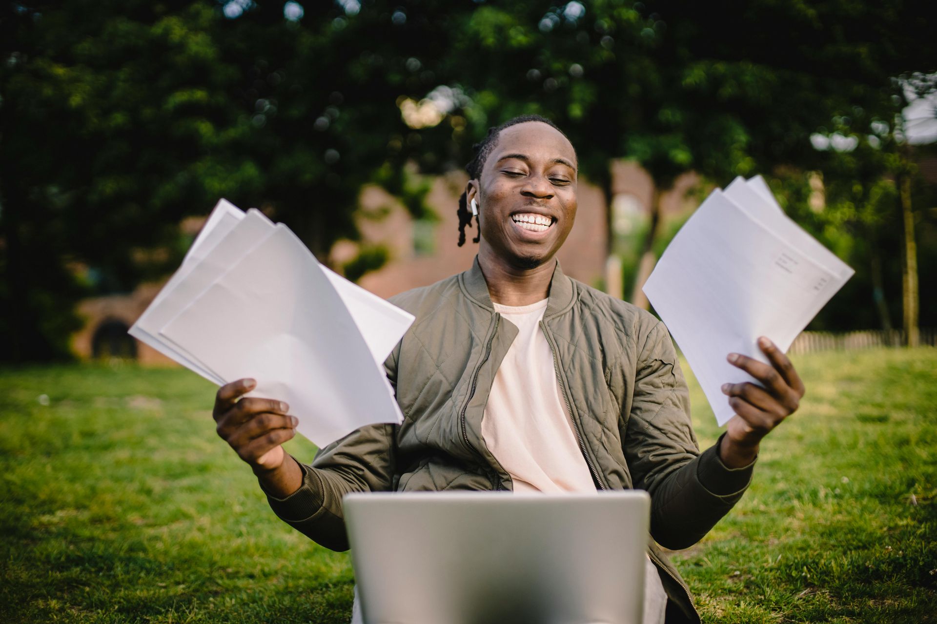 Man holding papers, smiling, laptop on lap, outside on grass.