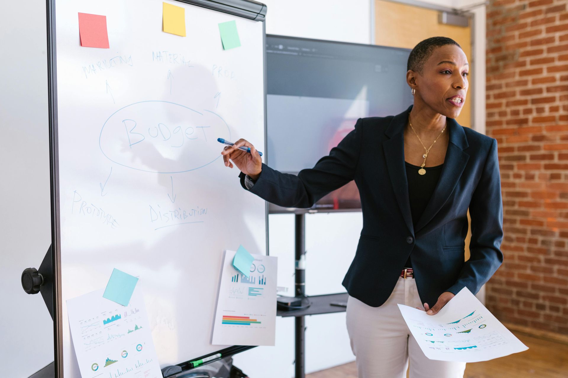 Woman in a blazer presenting at a whiteboard, pointing with a pen. Holding papers, a large screen behind.