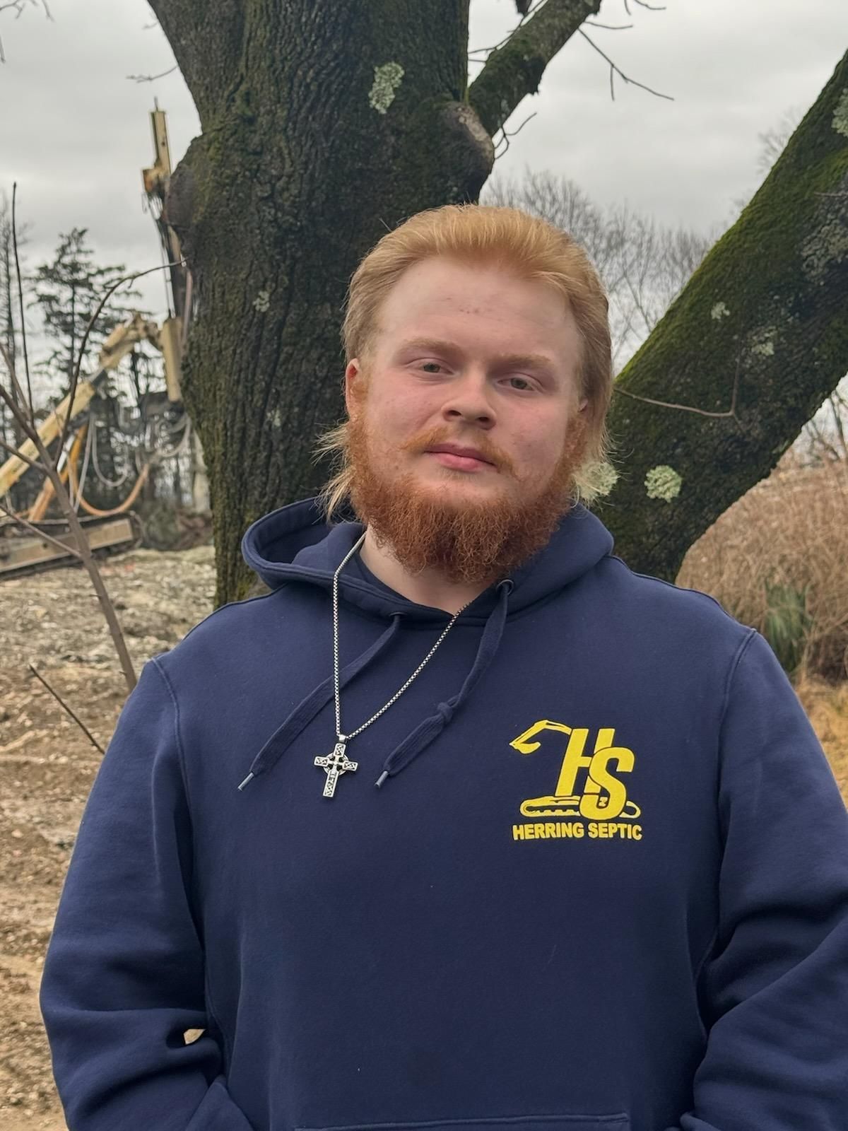 A man with a beard wearing a blue hoodie is standing in front of a tree.