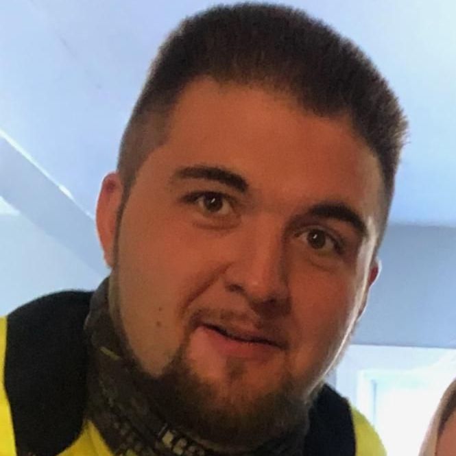 A man with a beard is wearing a yellow shirt and a black scarf around his neck.