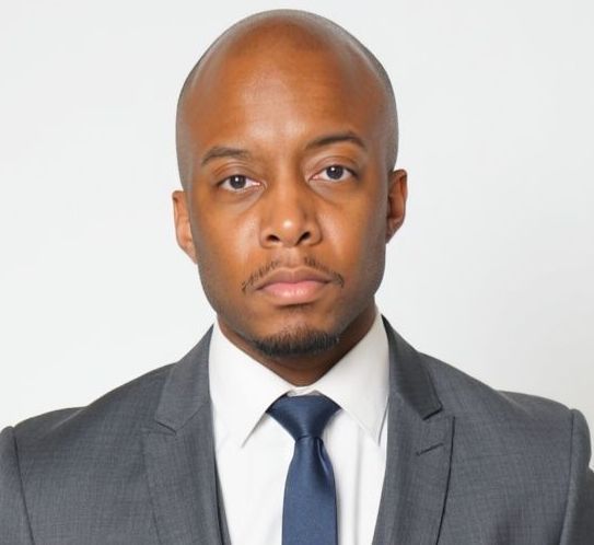 A Black man in a grey suit and tie looks directly at the camera, studio backdrop.