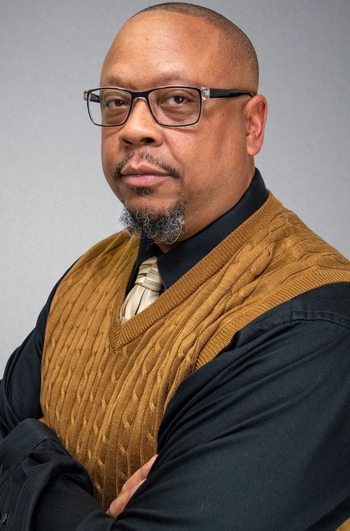 Man with glasses, arms crossed, wearing a black shirt, gold sweater vest, and tie; serious expression.