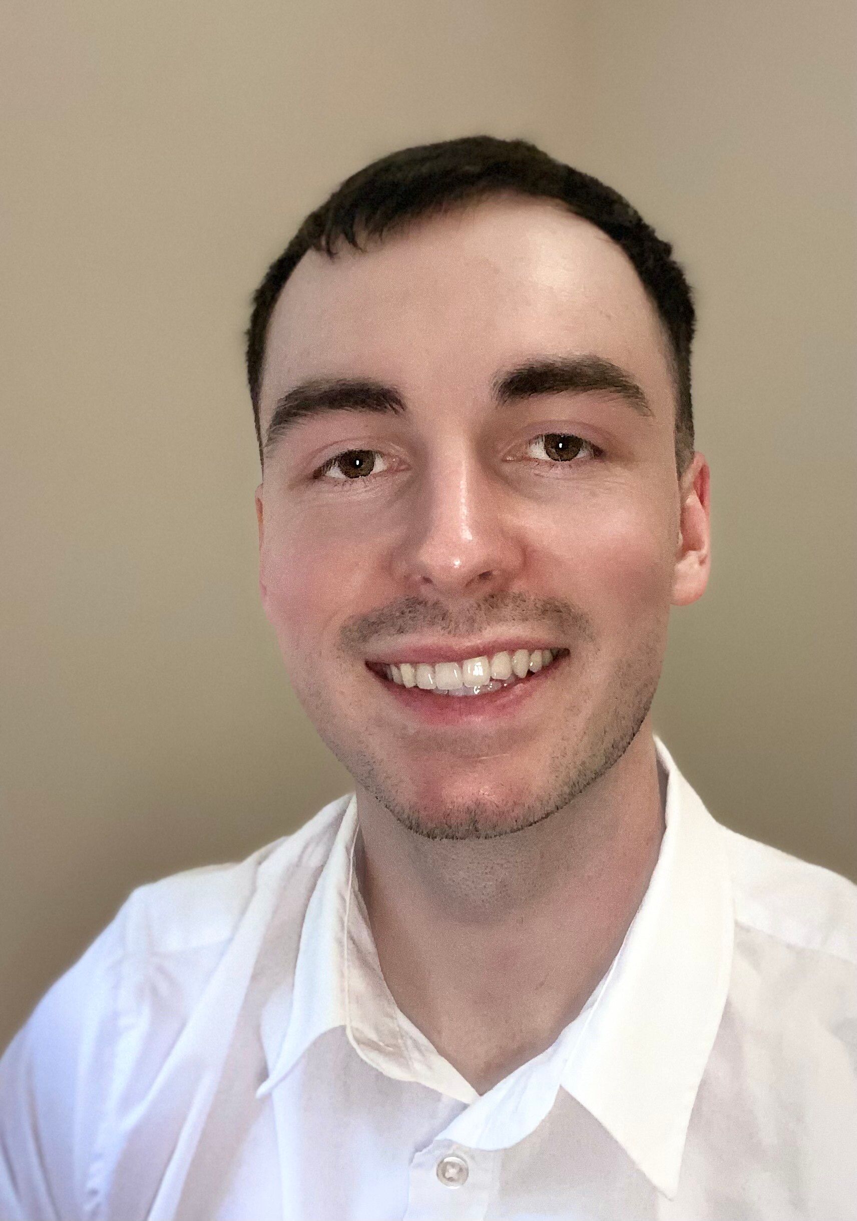 Man with short brown hair smiles, wearing a white shirt against a neutral wall.