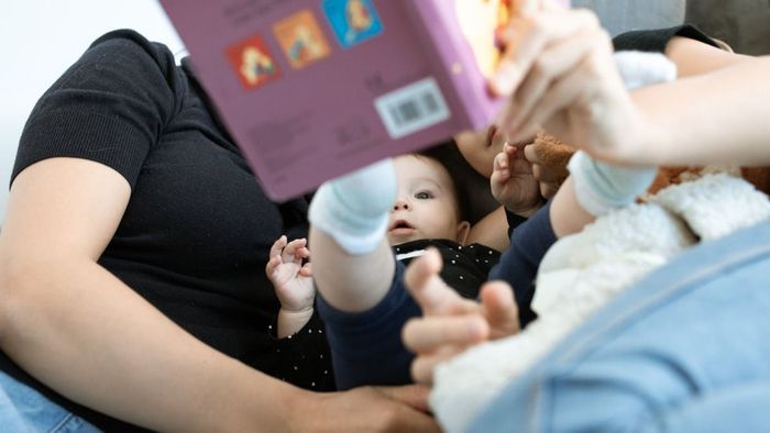 Un bebé acostado entre dos adultos, mirando un libro sostenido en alto, en el interior, con piel clara.