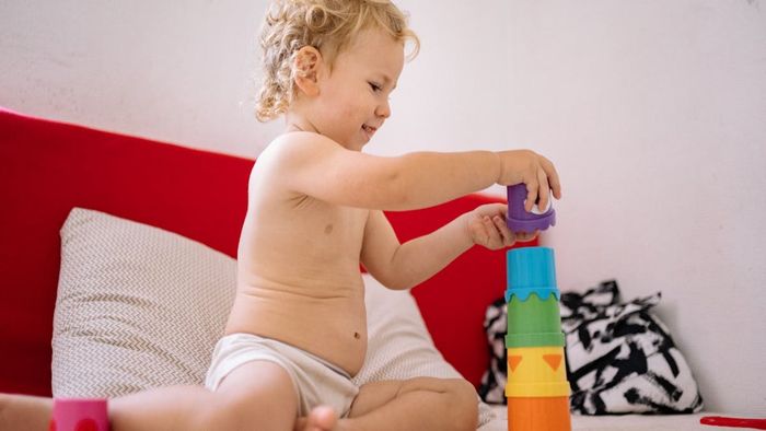 Un niño pequeño construye una torre con vasos coloridos apilables en el interior; una cabecera roja en el fondo.