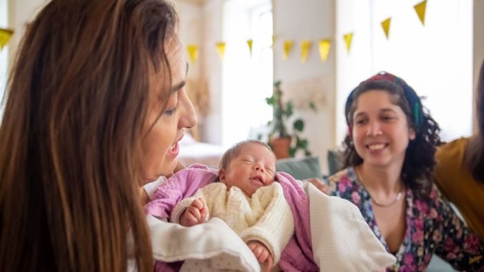 Mujer con un bebé recién nacido, sonriendo. Otra mujer sonríe al fondo. Colores pastel, interior.