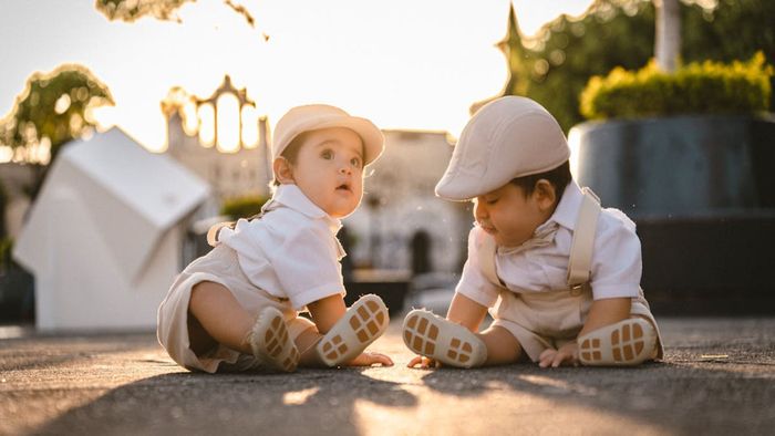 Dos bebés con ropa a juego, con sombreros, sentados en la acera, mirándose a la luz del sol.