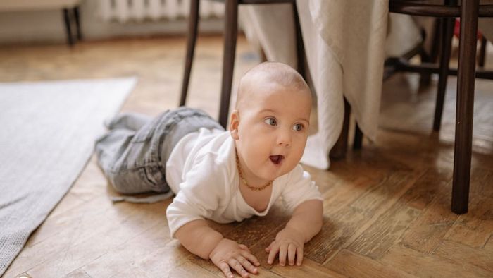 Bebé gateando sobre un piso de madera, vistiendo camisa blanca y jeans, luciendo sorprendido.