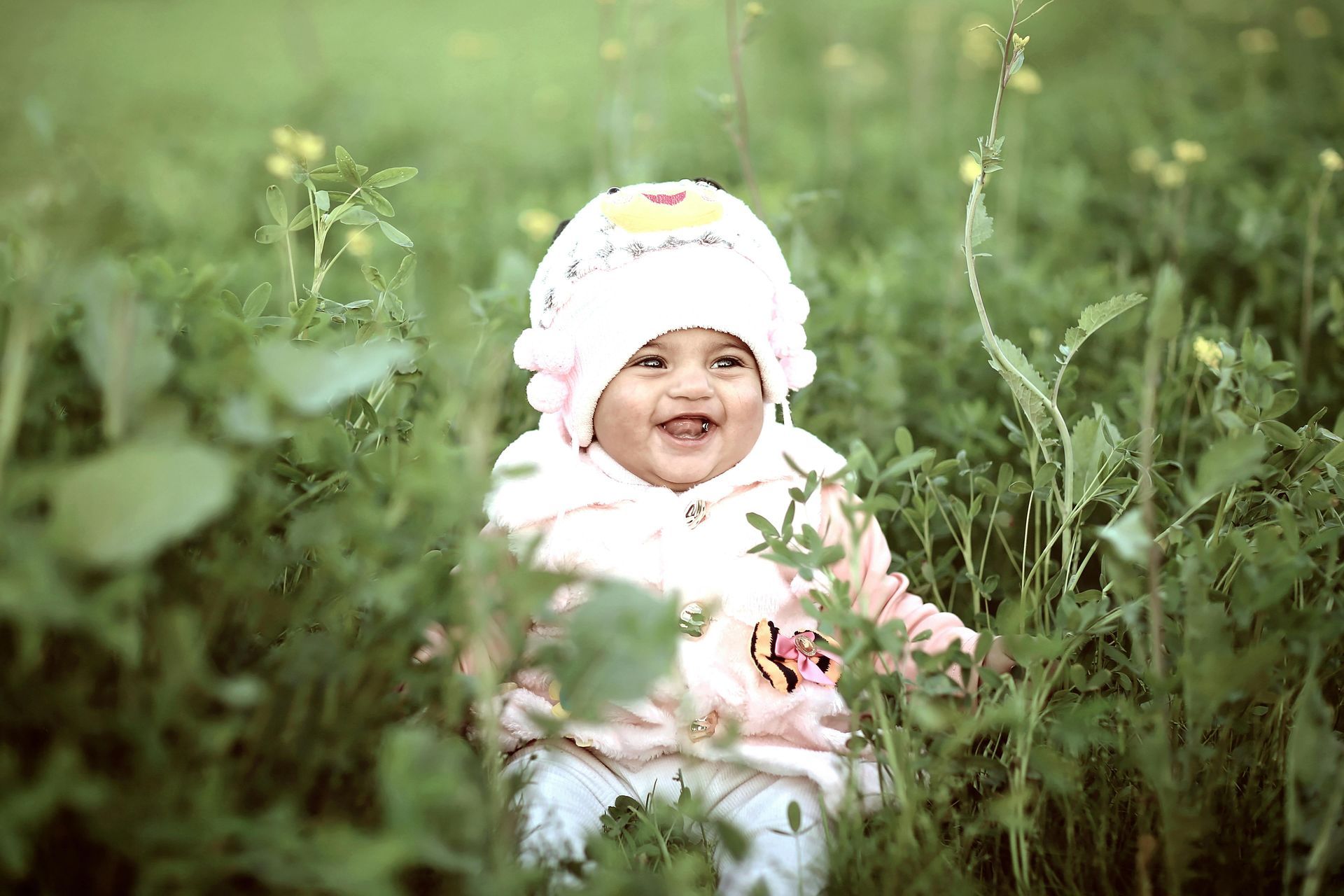 Un bebé sonriente con un sombrero y un suéter blancos se sienta en el césped verde.