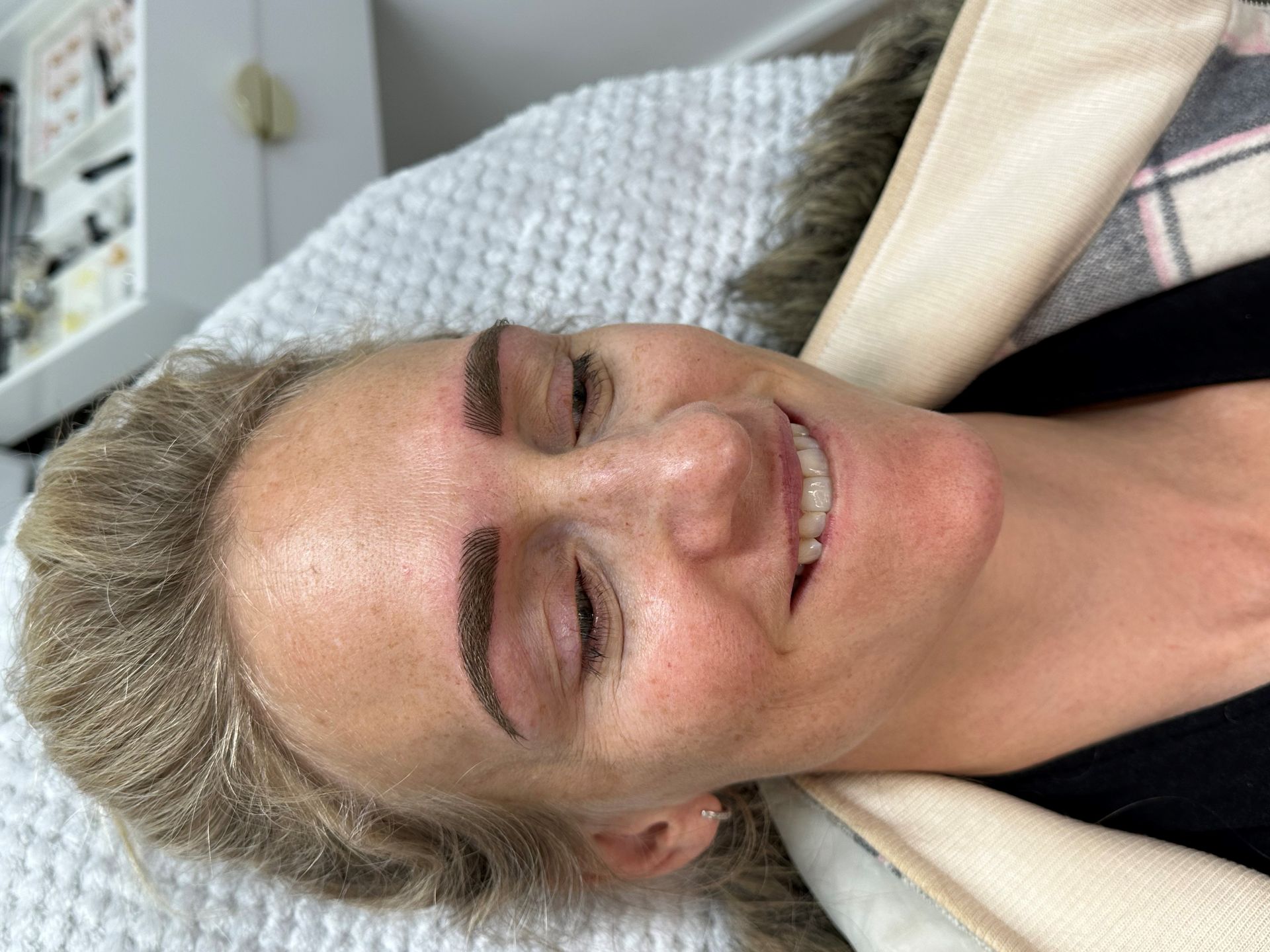 Woman with new eyebrow tattoos, smiling, lying on a bed with a fluffy blanket.